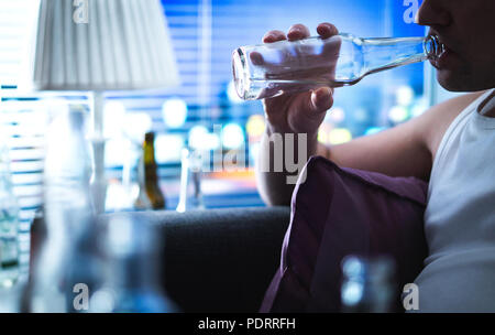Bevande alcoliche sorseggiando una bottiglia. L'alcolismo e l'abuso di alcool concetto. Drunken uomo seduto a casa sul lettino. Disoccupati triste ragazzo beve sul divano. Foto Stock