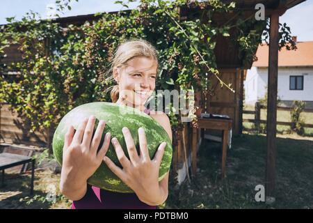 Ragazza ridere holding anguria pesante. Estate sul giardino sul retro della casa. Foto Stock
