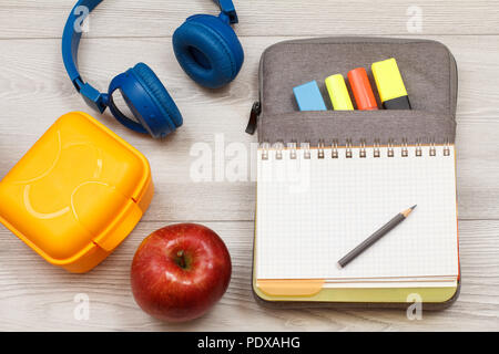 Forniture scolastiche. Giallo casella sandwich, Apple, cuffie, aprire il quaderno sul sacchetto custodia a matita con colore pennarelli e matita su grigio di tavole di legno. Foto Stock