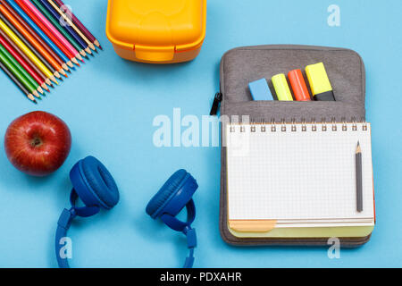 Apple, matite colorate, scatola di pranzo, cuffie, aprire il quaderno sul sacchetto custodia a matita con colore pennarelli e un marcatore su sfondo blu. Vista dall'alto. Indietro Foto Stock