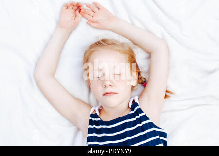 Closeup ritratto di carino redheaded adorabili bambini caucasici ragazza in striped nautico camicia senza maniche dormendo. Kid dreaming giacente su coltre bianca. Foto Stock