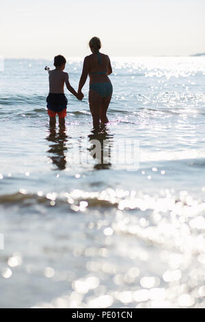 Madre e figlio divertirsi saltando le onde del mare nelle prime ore della sera mentre il sole tramonta ed è luccicante sulla pura acqua blu Foto Stock
