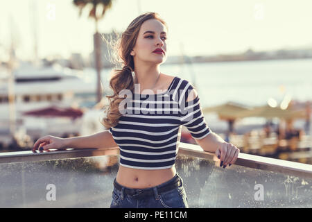 Donna bionda, modello di moda, in piedi in un porto con barche e mare sullo sfondo. Giovane e bella ragazza a strisce che indossa la t-shirt e jeans blu Foto Stock