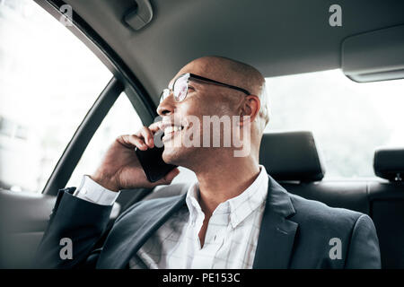 Imprenditore sorridente indossando occhiali parlando su telefono cellulare seduta nel suo sedan guardando fuori dalla finestra. L uomo nella gestione di aziende sul telefono cellulare sitt Foto Stock