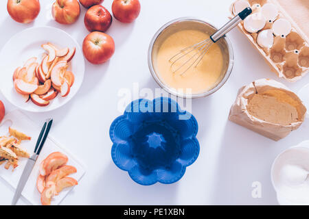 Ingredienti e strumenti per cucinare la torta di mele, vista dall'alto sulla cucina tavola bianco. Forma di silicone accanto al recipiente metallico riempito con l'impasto, frusta e tagliare la frutta, Foto Stock