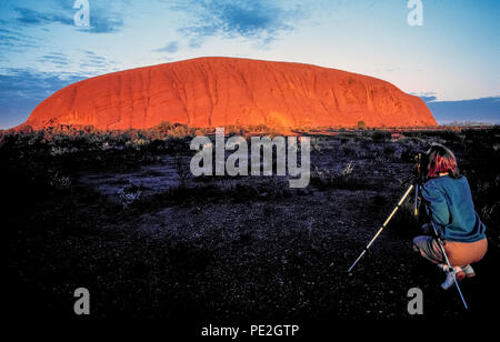 Un visitatore femmina fotografie alba a Uluru che provoca la terracotta-superficie colorata di questo mondo-famoso monolito di pietra arenaria a comparire in rosso fiammante luce in mezzo la outback scrublands del Territorio del Nord in Australia. Noto anche come Ayers Rock, la massiccia landmark sorge a circa 350 metri (1148 piedi) ) al di sopra del suolo e coperchi 8,6 chilometri quadrati (3.32 miglia quadrate) in Uluru-Kata Tjuta National Park. Si tratta di un luogo sacro per la zona indigene tribù aborigena, la Anangu, che ha chiamato le antiche rocce Uluru, il significato di "Terra Madre". Foto Stock