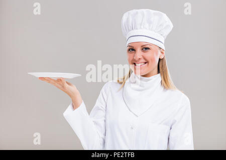 Bellissima femmina chef è tenendo la piastra su sfondo grigio. Foto Stock