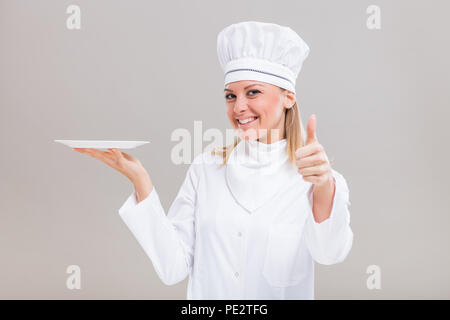 Bellissima femmina chef è la piastra di contenimento e mostrando il pollice su sfondo grigio. Foto Stock