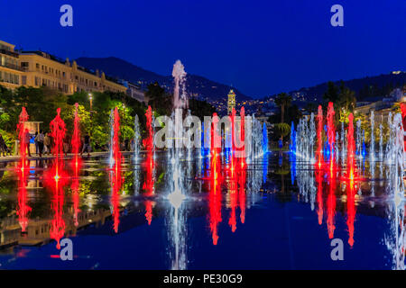 Fontana riflettente sulla Promenade du Paillon, circondato da un verde parco urbano, illuminato con il rosso e il blu delle luci al tramonto a Place Massena Massena o s Foto Stock