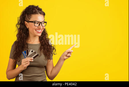 Contenuto giovane donna in bicchieri holding mucchio di carte di credito e rivolto lontano con il dito che mostra la vendita su sfondo giallo Foto Stock