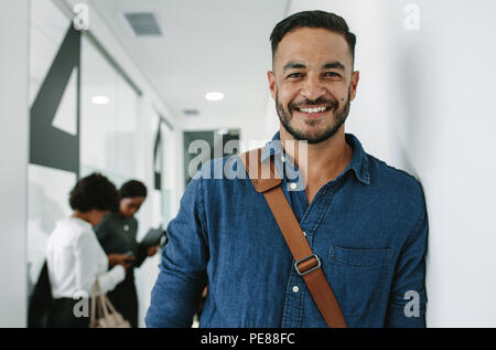 Ritratto di giovane casuals permanente al corridoio di office con i colleghi in background. Executive maschio appoggiata ad una parete in office corridoio. Foto Stock