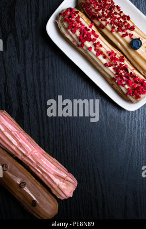 Deliziosi eclairs colorate con i chicchi di caffè, mirtillo, lampone su tavoli in legno nero Foto Stock