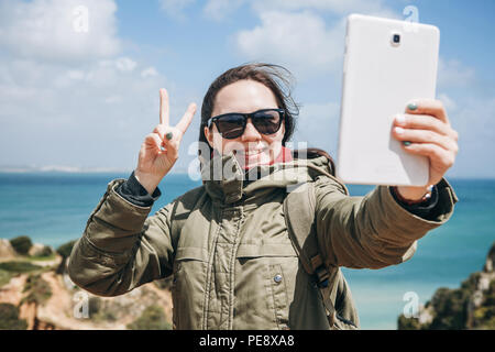 Una giovane donna turistico o blogger comunica sul tablet tramite Internet o storie i suoi abbonati su ciò che sta accadendo e mostra un segno con la mano. Foto Stock