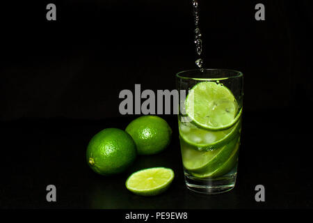 Estate rinfrescanti bevande e concetto. Torrente versa in un bicchiere di bibita con ghiaccio e fresche e mature green slice limes su sfondo nero. Homema Foto Stock