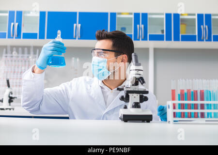 Duro lavoro. Professional attraente laboratorian maschio lampadina in salita mentre lo sta guardando e indossare occhiali di sicurezza e la maschera Foto Stock