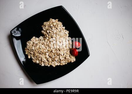 Vista superiore fiocchi di avena sotto forma di un cuore su una piastra nera e fragole fresche Foto Stock