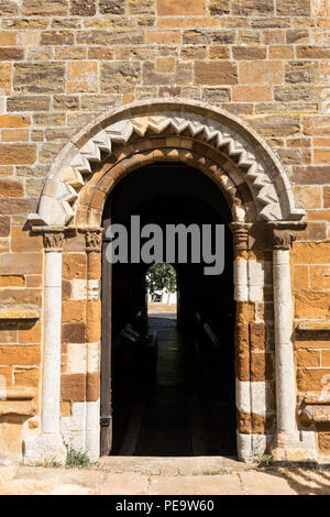 Spratton, Northamptonshire, 24-07-18. Una porta aperta in una parete è sormontato da un scolpito Arco Normanno. All'interno è buio e una porta che lasciano entrare la luce. Foto Stock