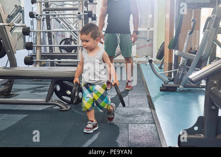 Piccolo figlio di aiutare il padre sulla formazione in palestra con le pesanti manubri al simulatore metallico. Padre a piedi dietro. Il mantenimento di uno stile di vita sano, sportivo, montare il corpo, forti muscoli, tempo per la famiglia. Foto Stock