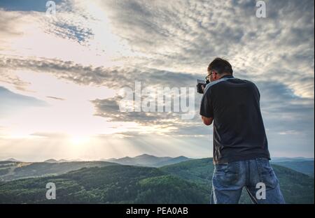 Fotografo di natura creare arte sul punto di vista nelle montagne. Uomo che guarda nel mirino del suo grande fotocamera, prendendo foto di borning giornata incredibile. Foto Stock