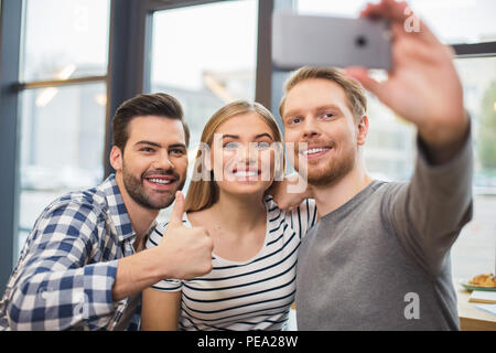 E ora sorriso. Bella piacevole lieto uomo in piedi insieme con i suoi amici e tenendo lo smartphone tenendo selfie Foto Stock