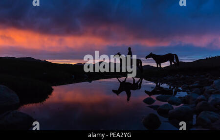 Un cavaliere nomade conduce un cavallo su acqua nel deserto mongolo sotto un tramonto spettacolare. Foto Stock