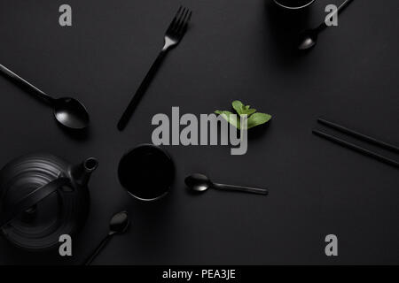 Vista superiore della teiera nero, bastoncini, vari cucchiai, forcella, Cup e le foglie di menta sulla tavola nera Foto Stock
