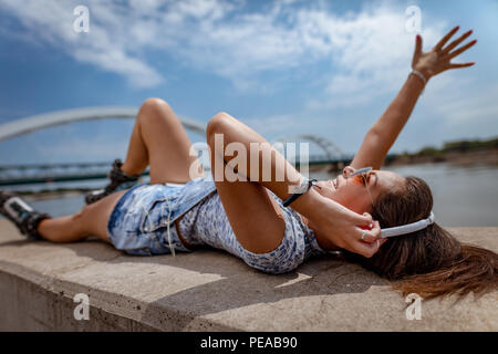 Carino sorridente giovane donna con pattini a rotelle ascoltando musica dal tuo telefono e godendo della città riverbank presso la bella giornata d'estate. Foto Stock