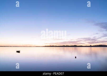 Piccolo dinghy sfocato oscillante su acqua nella lunga esposizione al di sopra di Tauranga Harbour sunrise si illumina in gamma di blu,golden e sfumature di rosa in tutta l'acqua all'alba Foto Stock