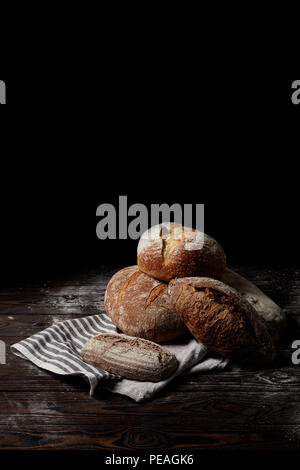 Vista ingrandita di vari tipi di pane e il sacco sul tavolo in legno di copertura mediante la farina isolato su sfondo nero Foto Stock