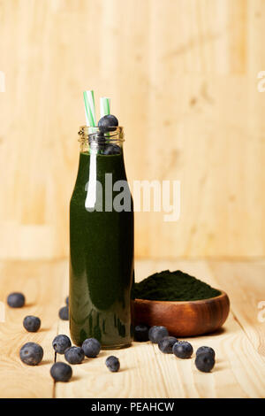 Ciotola con polvere spiruluna, bottiglia di spirulina smoothie con mirtilli e cannuccia sul tavolo di legno Foto Stock