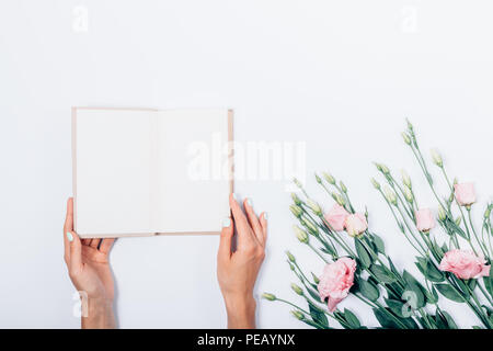 La femmina mani tenendo aperto il libro vicino ad un mazzo di fiori di colore rosa su sfondo bianco, vista dall'alto. La donna che mostra le pagine bianche di un libro di testo, piatto laici. Foto Stock