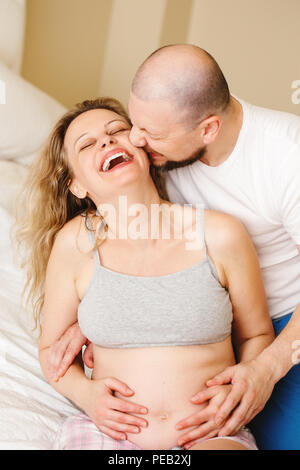 Ritratto di sorridere ridere bianchi giovani caucasici la mezza età matura, donna incinta con il marito in camera sul lettino abbracciando cuddling, lifestyle materni Foto Stock