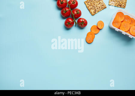 Vista superiore della disposizione del contenitore per alimenti con freschi fette di carote, pomodori e cookie isolato su blu Foto Stock