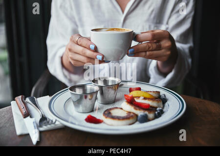 Ritagliato colpo di donna azienda tazza di caffè presso il ristorante sopra il formaggio frittelle sul tavolo Foto Stock