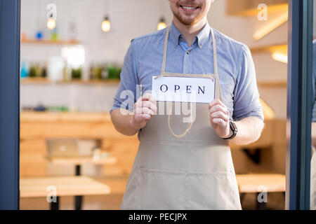 Tempo per essere aperto. Positivo allegro giovane uomo in piedi dietro la porta di vetro e mantenere l'etichetta etichetta mentre mostra che il Cafe è aperto Foto Stock