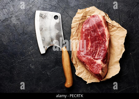 Crudo fresco di carne in marmo nero bistecca angus su carta marrone e carne cleaver. Carne su sfondo nero con copia spazio. Vista dall'alto. Foto Stock