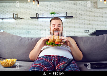 Fat funny man in pigiama mangiare un hamburger sul divano di casa. Foto Stock