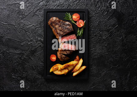 Carne Picanha bistecca, tradizionale taglio brasiliano con pomodori, spicchi di patate e rosmarino carne nero tagliere con copia spazio. Nero lo sfondo di calcestruzzo. Foto Stock