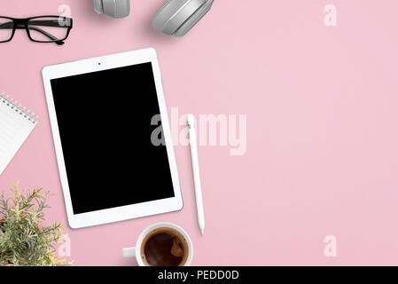 Tablet mockup in rosa scrivania circondata con le cuffie, bicchieri, notepad, impianti, una tazza di caffè e penna. Spazio libero accanto per il testo. Lay piatto compo Foto Stock