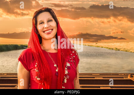 Donna mediterranea indossa velo rosso e sorridente sul fiume sfondo Foto Stock