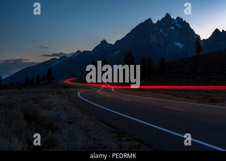 Luci di coda in autostrada di notte al Parco Nazionale di Grand Teton con sfondo della gamma della montagna e il sole che tramonta dietro. Foto Stock