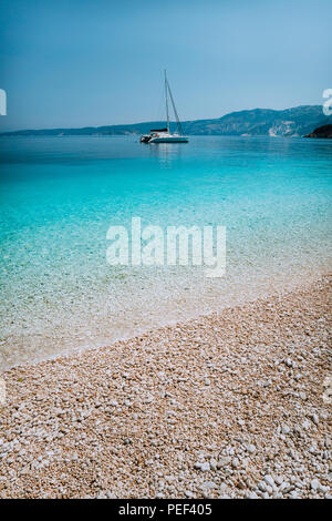 Spiaggia ghiaiosa con chiara pura acqua azzurra e catamarano a vela yacht barca ad ancorare su orizzonte Foto Stock