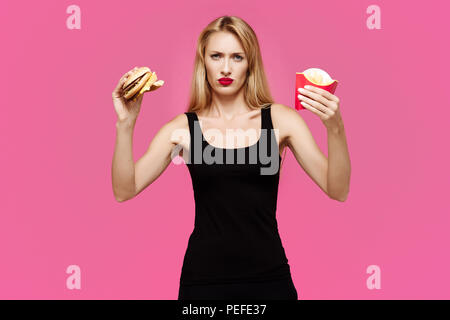 Giovane bella ragazza slanciata su un sfondo rosa detiene un hamburger e patatine nelle sue mani. Concetto di junk food Foto Stock