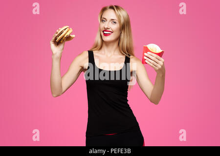 Giovane bella ragazza slanciata con sorriso bianco su uno sfondo rosa detiene un hamburger e patatine nelle sue mani e sorridente. Concetto di junk food Foto Stock