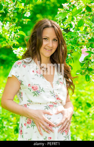 Ritratto di un felice donna incinta in una fioritura d'estate il parco Foto Stock