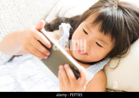 Carino bambina divertirsi a giocare con il gioco su smart phone sdraiato sul divano. Foto Stock