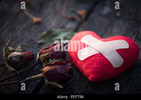 Aiuti di prima fascia imbottita e striscia di gesso Patch medico incollato sul cuore rosso con secchi rosa rossa su sfondo di legno. Cuore rotto, amore e San Valentino da Foto Stock