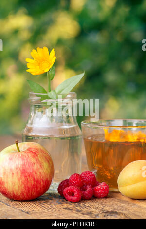 Frutti maturi di mela, albicocca e lamponi, calendula fiore con uno stelo in un vasetto di vetro e la tazza di tè verde su assi di legno con naturale sfocata Foto Stock