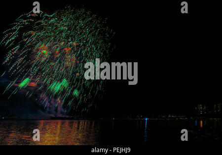 Multicolore di fuochi d'artificio sull'oceano nella grande città, sagome di gente sulla spiaggia di moderni edifici sullo sfondo, incandescente e riflettendo Foto Stock