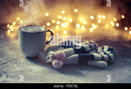 Atmosfera accogliente con l'inverno di caffè o di tè la tazza con il vapore, guanti di lana su luci bokeh di fondo Foto Stock